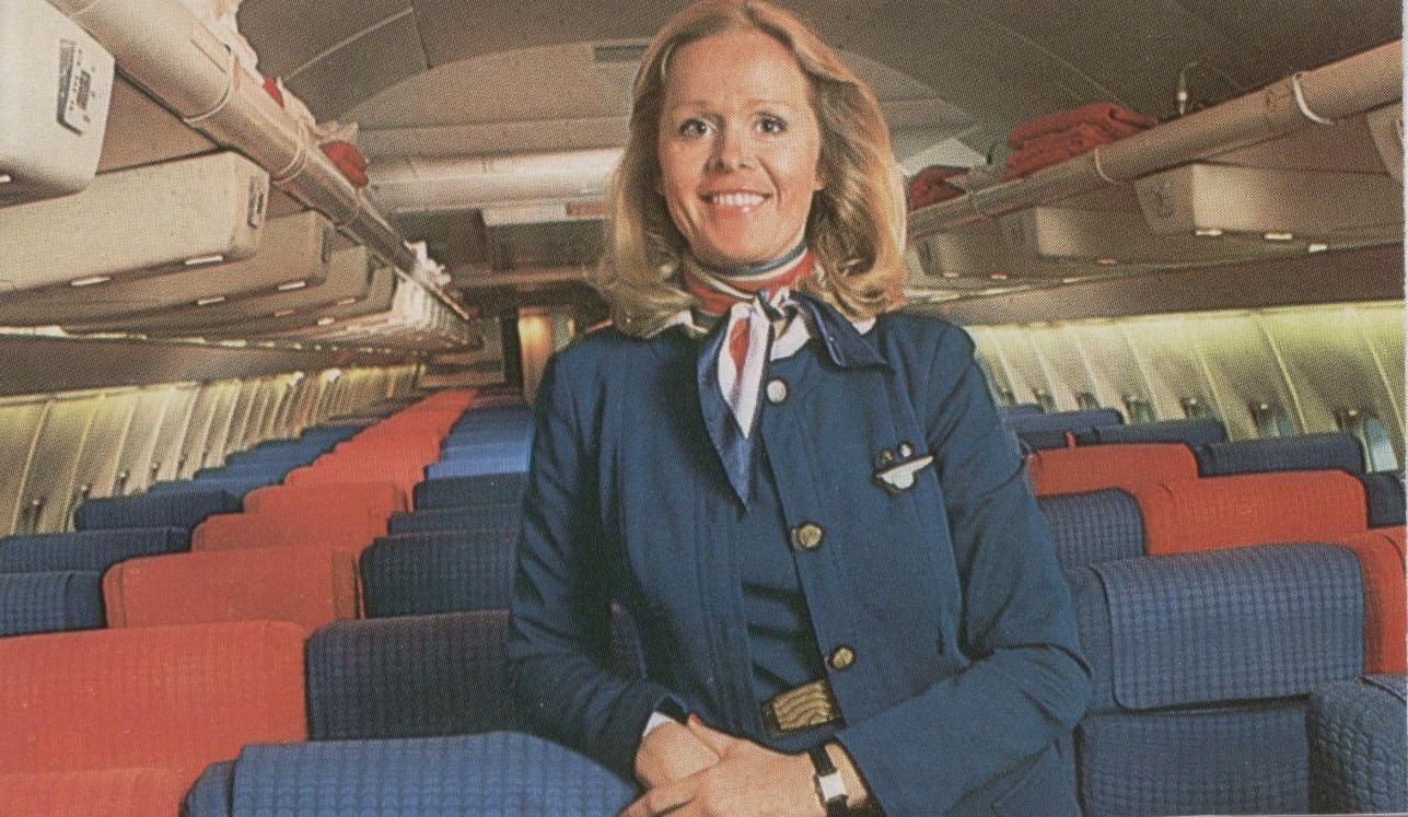 1970s A Flight Attendant in a uniform designed by Edith Head stands in the aisle of a Pan Am 707 reconfigured for charter service with 180 economy seats.
